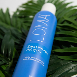 A blue bottle of LOMA Extra Firm Hold Hairspray set against a backdrop of green leaves.