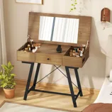 A wooden vanity table with a flip-up mirror, featuring black metal legs, is organized with various cosmetics, a phone stand, and a small potted plant nearby.