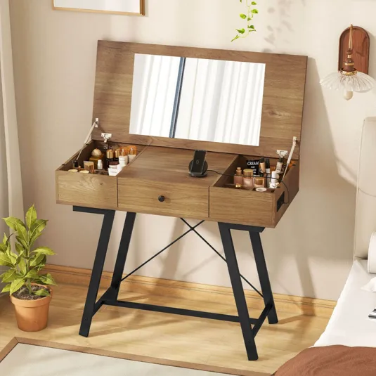 A wooden vanity table with a flip-up mirror, featuring black metal legs, is organized with various cosmetics, a phone stand, and a small potted plant nearby.