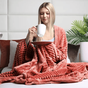 A person sits on a bed, wrapped in an oversized, textured, coral-colored corduroy plush throw blanket, holding a white mug on a plate. A potted plant is visible in the background, adding a cozy touch to the scene.