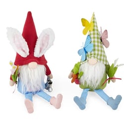 Two Easter gnomes with white beards and soft fabric hats, one red with bunny ears, holding a lantern, and the other green plaid with butterflies and flowers. Each wears blue pants and has decorative accents like carrots and more flowers.