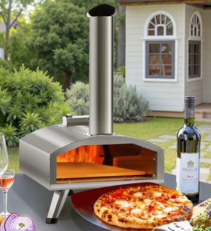 A stainless steel wood-burning pizza oven with a chimney, positioned outdoors on a table beside a cooked pizza, a wine bottle, a wine glass, and sliced onions, with a garden setting and a small white building in the background.