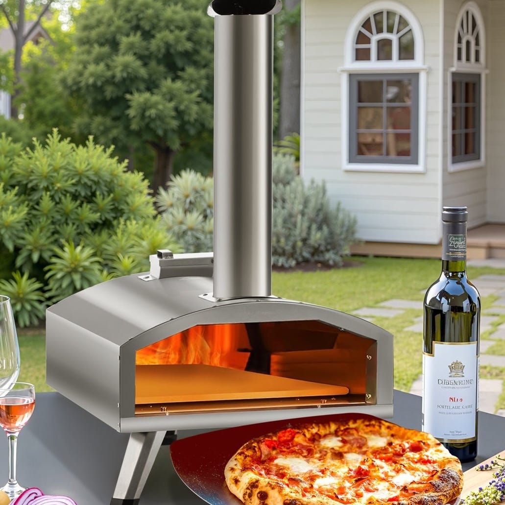 A stainless steel wood-burning pizza oven with a chimney, positioned outdoors on a table beside a cooked pizza, a wine bottle, a wine glass, and sliced onions, with a garden setting and a small white building in the background.