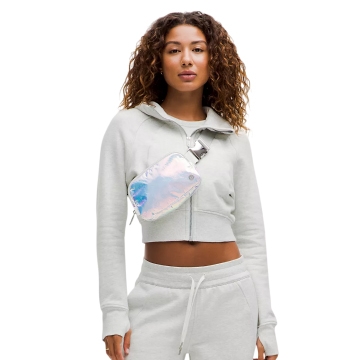 A woman wears a lululemon iridescent belt bag over a light grey hoodie and sweatpants. The bag has a shiny, multicolored surface and a front zip closure, styled as a crossbody accessory. She has curly brown hair.