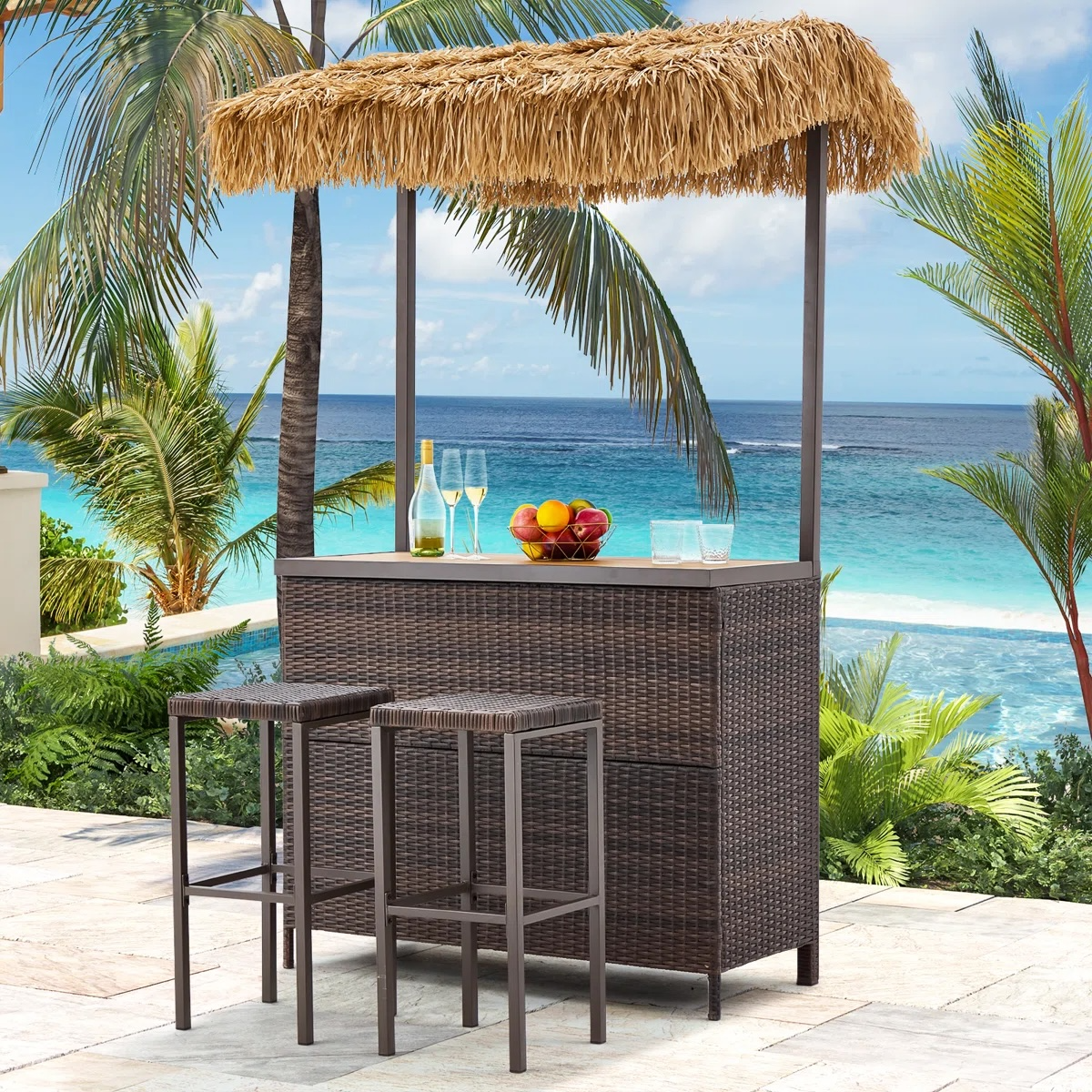 Outdoor tiki bar set with a thatched roof, two bar stools, and a wicker-style design, set against a tropical ocean backdrop. Includes a fruit bowl, wine bottle, and glasses on the countertop.