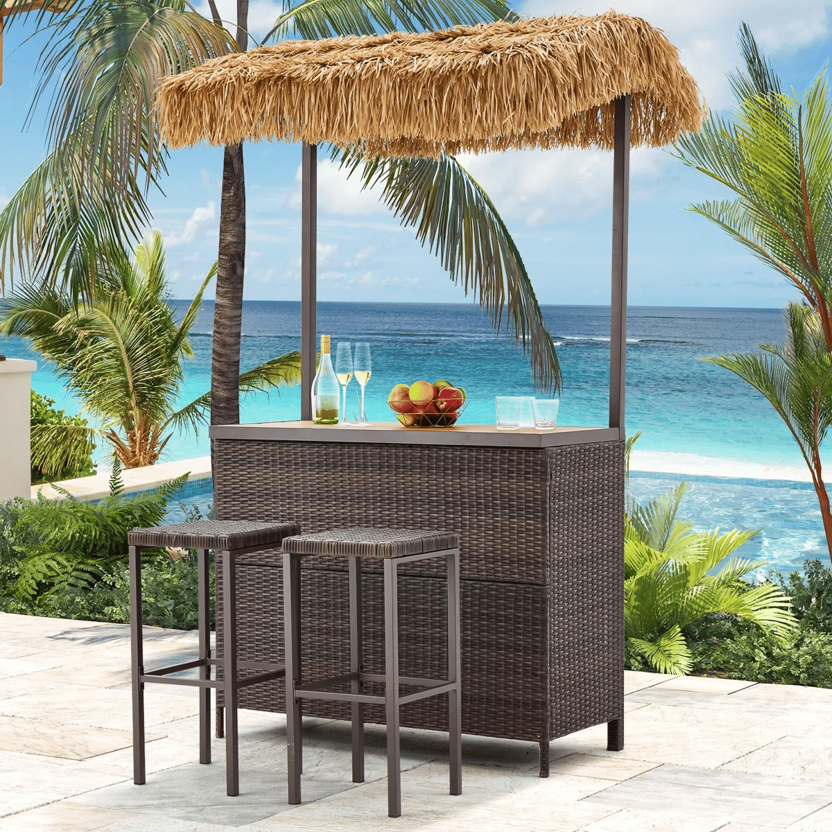 Outdoor tiki bar set with a thatched roof, two bar stools, and a wicker-style design, set against a tropical ocean backdrop. Includes a fruit bowl, wine bottle, and glasses on the countertop.
