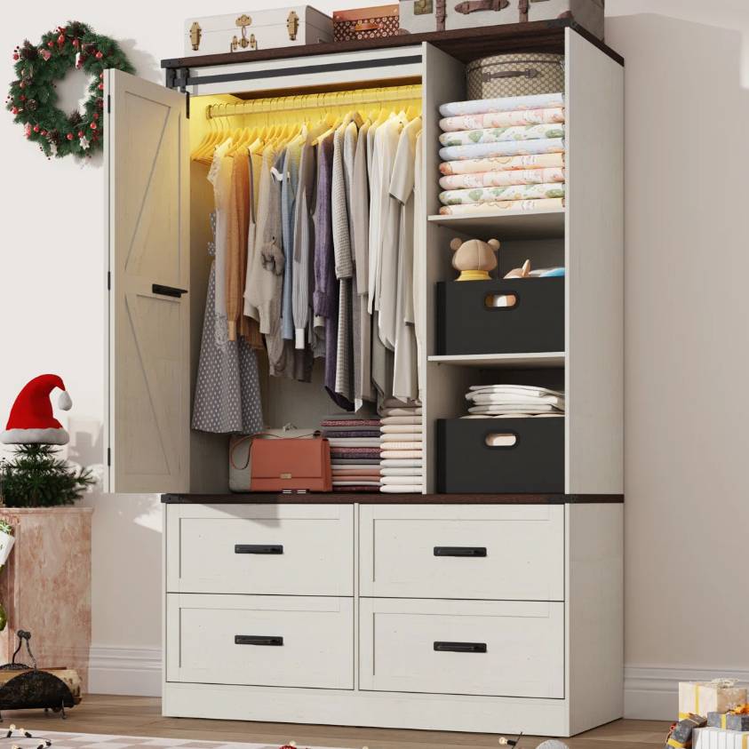 The August Grove Armoire is a white wooden wardrobe with a sliding barn door and includes a hanging section with LED lights, shelves for folded clothes, decorative boxes, and four lower drawers. Top features luggage-style boxes, complemented by seasonal decor.