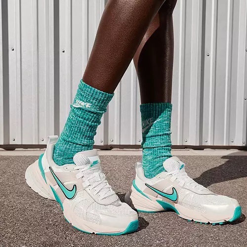 A pair of white sneakers with teal accents and teal Nike socks are shown on a person's feet against a corrugated wall background.