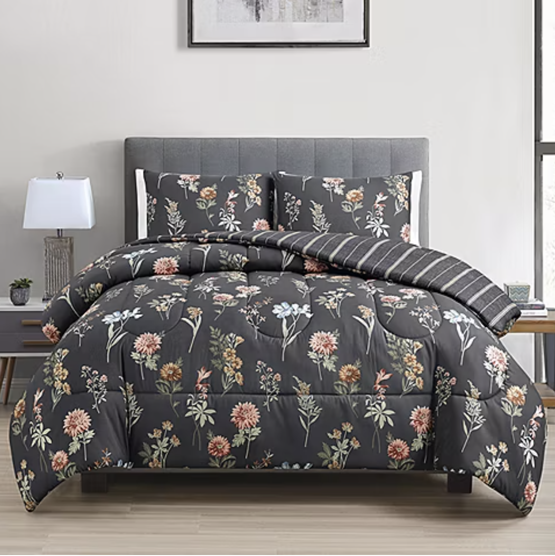 A bed features a floral-patterned comforter set with matching pillow shams in dark gray tones, complemented by a striped throw. The scene includes a side table with a lamp and a small plant, set in a modern bedroom.