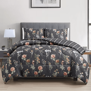 A bed features a floral-patterned comforter set with matching pillow shams in dark gray tones, complemented by a striped throw. The scene includes a side table with a lamp and a small plant, set in a modern bedroom.