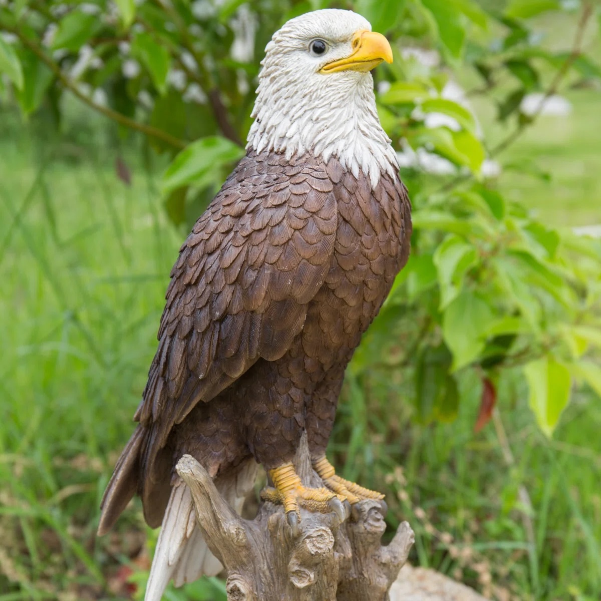 A realistic bald eagle sculpture with detailed feathers and a striking yellow beak, perched on a branch, set against a natural green background.