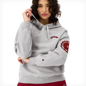 A person wearing a gray Champion hoodie with red logo details on the left chest and sleeve.