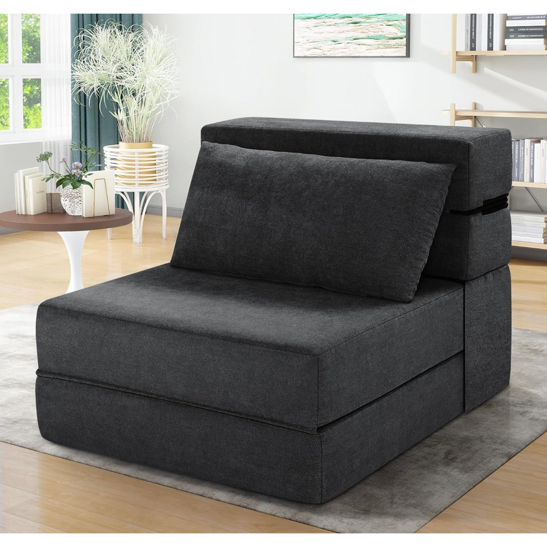 A dark gray convertible chair with a cushioned backrest and wide seat, positioned in a modern living room with books and plants nearby.