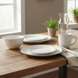 A dining set featuring white tableware, including a bowl, two plates, and a mug on a wooden table. A glass pitcher of water and a small potted plant are also present.