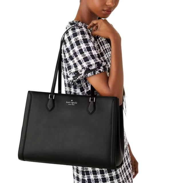 A person is holding a large black Kate Spade tote bag. The bag features a sleek design with two handles and a subtle logo at the top center.