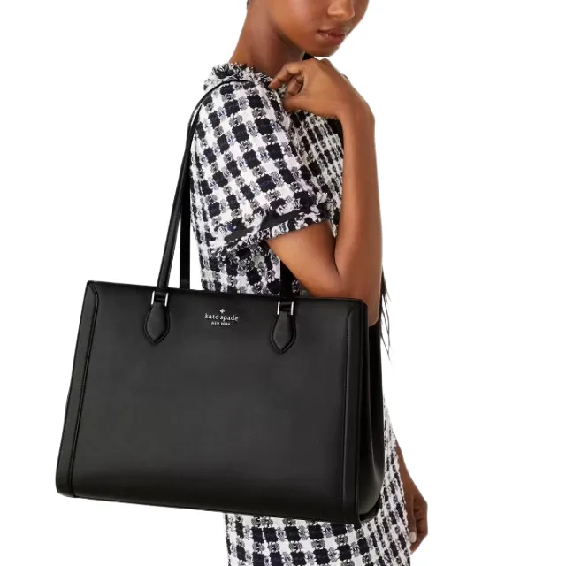 A person is holding a large black Kate Spade tote bag. The bag features a sleek design with two handles and a subtle logo at the top center.