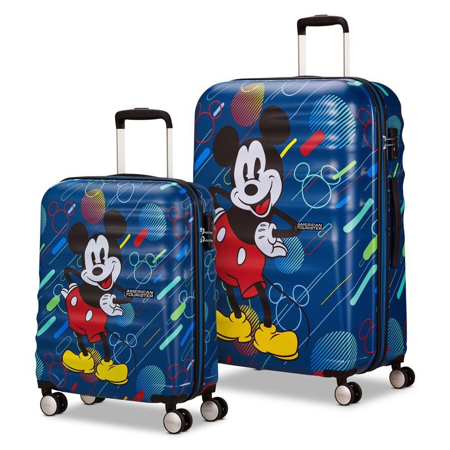 Two hard-shell suitcases feature a colorful design with a cartoon character. The smaller suitcase has four wheels, and the larger one is similar in style. Both are by American Tourister.