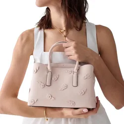 A person is holding a pink handbag with floral 3D embellishments. The handbag has a structured design, dual handles, and subtle branding.