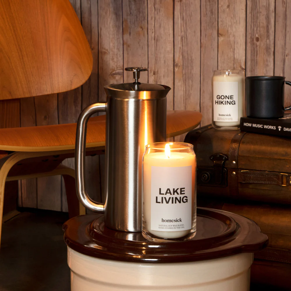 A lit \“LAKE LIVING\“ candle by Homesick next to a stainless steel French press on a ceramic stand, with a wooden chair and rustic backdrop.