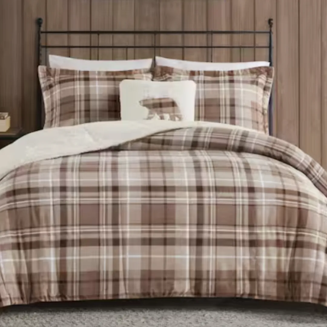 A bed features a plaid-patterned comforter set in brown tones with matching pillowcases and a small pillow with a bear design. The backdrop includes a wooden paneled wall.