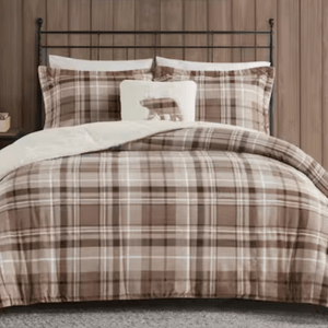 A bed features a plaid-patterned comforter set in brown tones with matching pillowcases and a small pillow with a bear design. The backdrop includes a wooden paneled wall.