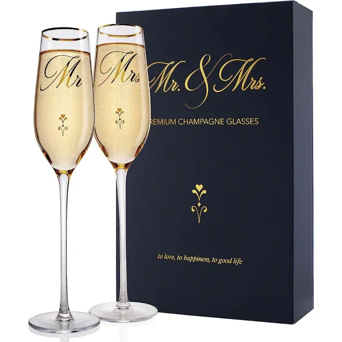 Two champagne flutes labeled \“Mr\“ and \“Mrs\“ are in front of a box that says \“Mr. & Mrs. Premium Champagne Glasses\“ and includes the phrase \“to love, to happiness, to good life.\“