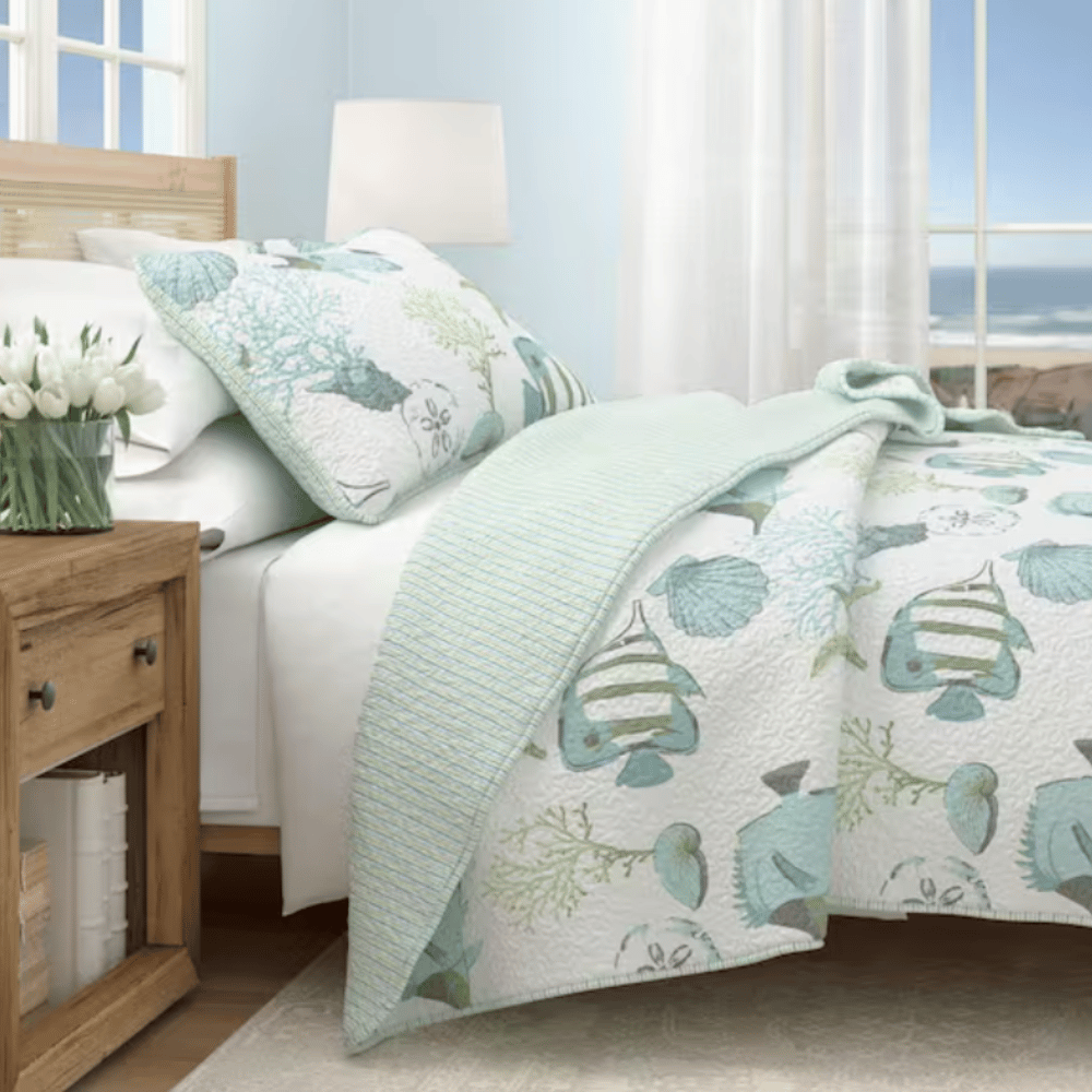 A coastal-themed bedroom setup featuring a bed with aqua and white bedding with fish and seashell motifs, complemented by a wooden nightstand with a vase of white tulips. A large window offers a view of the ocean.
