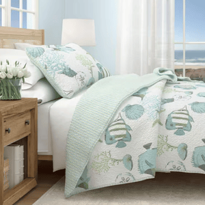 A coastal-themed bedroom setup featuring a bed with aqua and white bedding with fish and seashell motifs, complemented by a wooden nightstand with a vase of white tulips. A large window offers a view of the ocean.