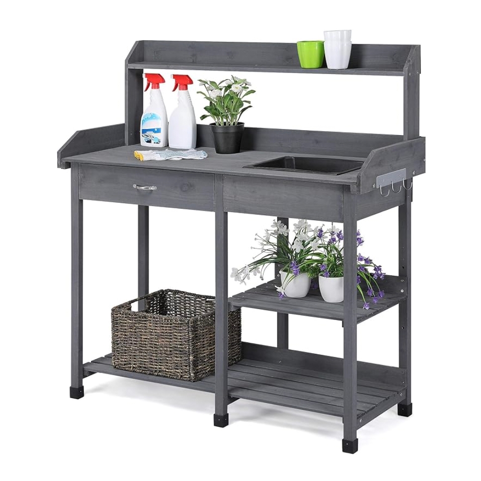 A gray wooden potting table features a workspace with a built-in sink, a top shelf with pots and spray bottles, a drawer, and side hooks. Two lower shelves hold a woven basket and potted plants.