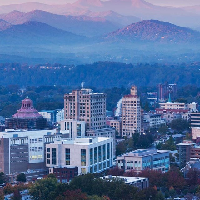 A skyline view of a city with modern and historic buildings in the foreground, surrounded by lush greenery and distant mountains under a hazy sky.