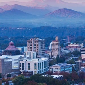 A skyline view of a city with modern and historic buildings in the foreground, surrounded by lush greenery and distant mountains under a hazy sky.