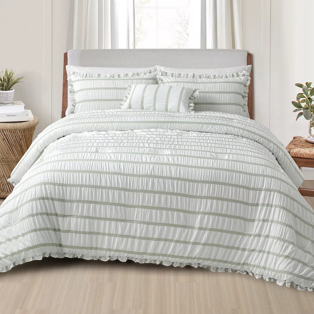 A bed features a ruffled white and light gray striped comforter set with matching pillow shams and a decorative pillow. The setting includes a wooden nightstand with a plant and books.