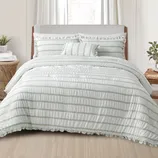 A bed features a ruffled white and light gray striped comforter set with matching pillow shams and a decorative pillow. The setting includes a wooden nightstand with a plant and books.