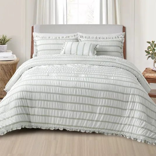 A bed features a ruffled white and light gray striped comforter set with matching pillow shams and a decorative pillow. The setting includes a wooden nightstand with a plant and books.