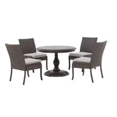 A round outdoor dining set with a central pedestal table and four wicker chairs featuring light cushions.