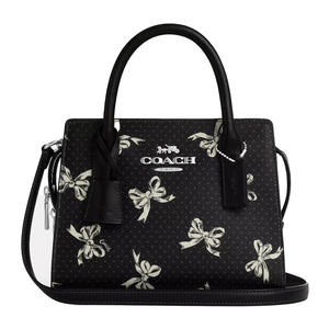 A black Coach Outlet handbag features a white bow print, silver hardware, and dual top handles with a detachable shoulder strap. The bag includes the Coach logo and a leather tag.