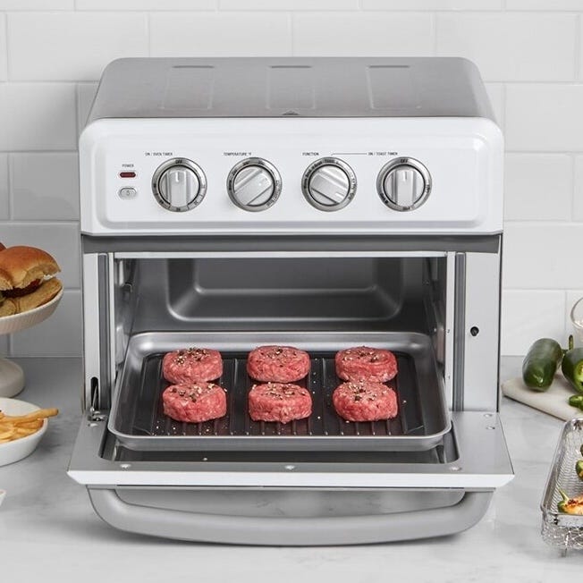 A countertop convection oven with a stainless steel finish is open, displaying a tray with six uncooked burger patties. The setting includes a plated hamburger and fries nearby, along with jalapeños.