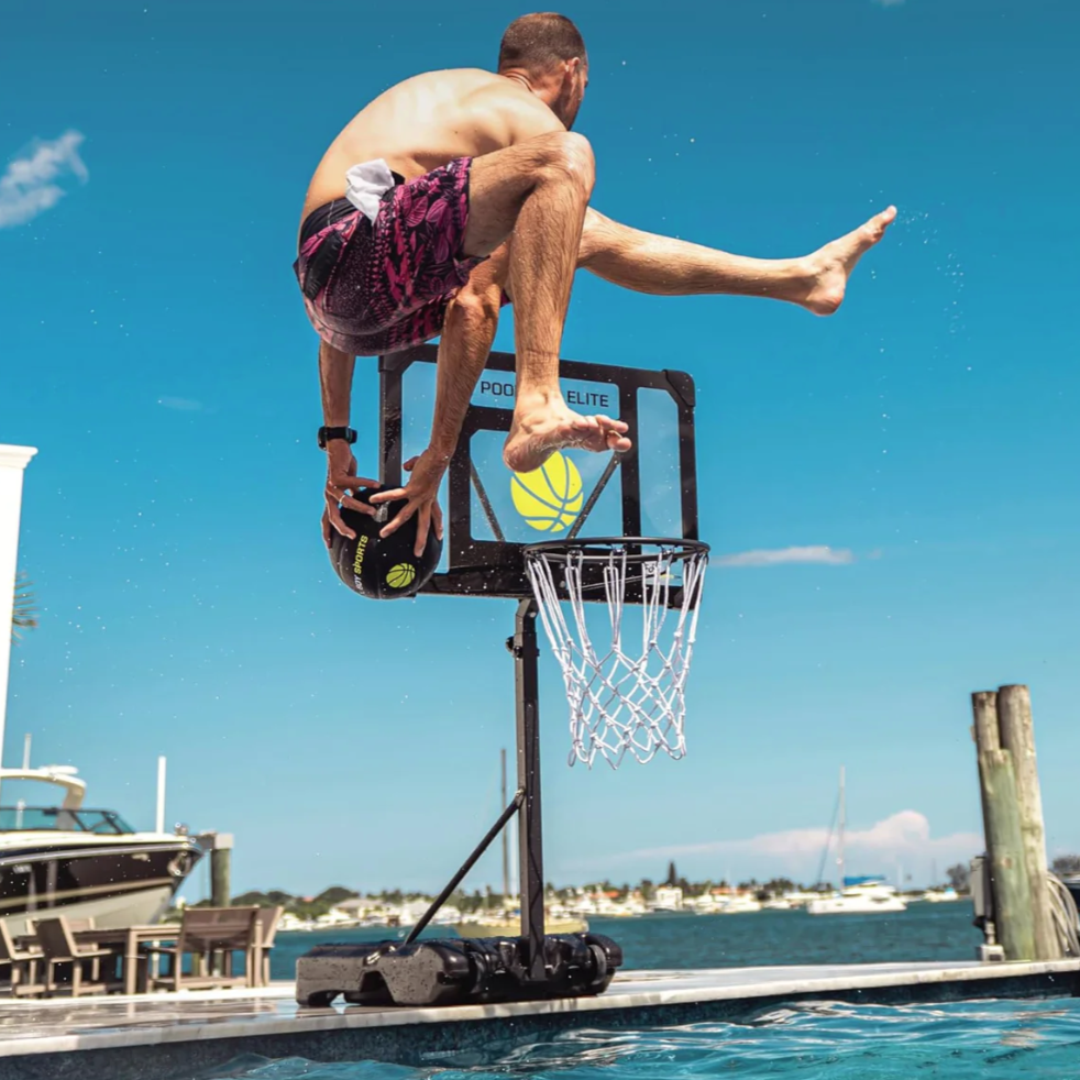 A person is performing a basketball trick on a hoop set up next to a pool, holding a basketball while jumping over the hoop.