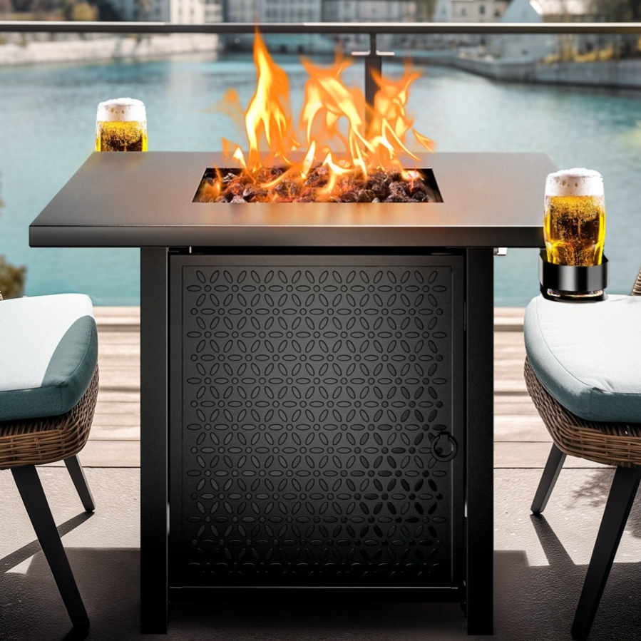 A square outdoor fire pit table with a decorative panel is set between two wicker chairs with blue cushions, overlooking a scenic body of water. Two glasses of beer are placed on the table's edges.
