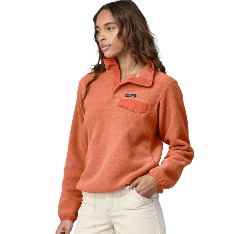 A woman is wearing an orange Patagonia fleece pullover with a snap-button collar and chest pocket, paired with light-colored pants.