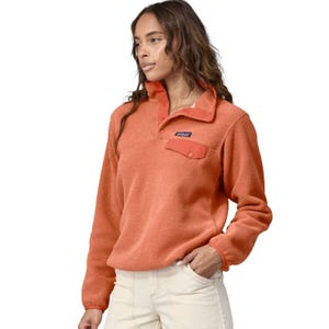 A woman is wearing an orange Patagonia fleece pullover with a snap-button collar and chest pocket, paired with light-colored pants.