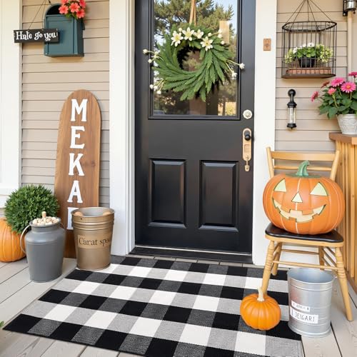 Front porch with black door, adorned with a wreath, features a jack-o'-lantern, pumpkins, a checkered rug, potted plants, and a \“Meka\“ sign.