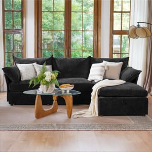 A dark gray sectional sofa with white pillows and a blanket is positioned in front of a large window. A glass coffee table with a wooden base holds a plant and a small tray with food.
