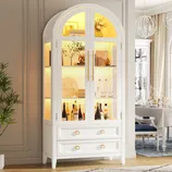 White cabinet with glass doors and interior lighting, featuring shelves with bottles, glassware, and decor items, plus two bottom drawers with gold handles.