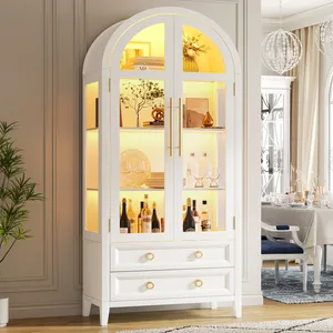 White cabinet with glass doors and interior lighting, featuring shelves with bottles, glassware, and decor items, plus two bottom drawers with gold handles.