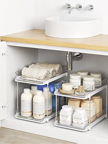 Two sliding metal shelf organizers containing toiletries are installed inside a cabinet beneath a sink with a wooden counter.