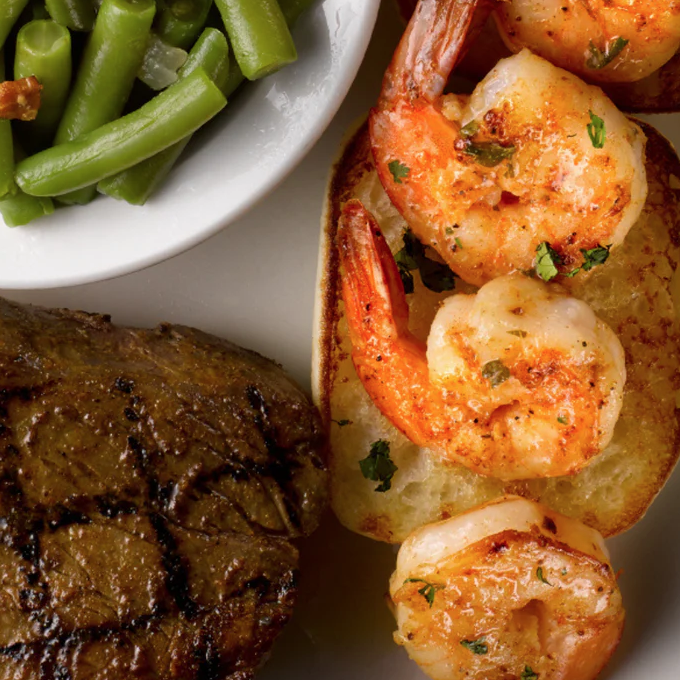 Grilled shrimp with herbs on toast, a steak with grill marks, and a side of green beans with bacon in a bowl.