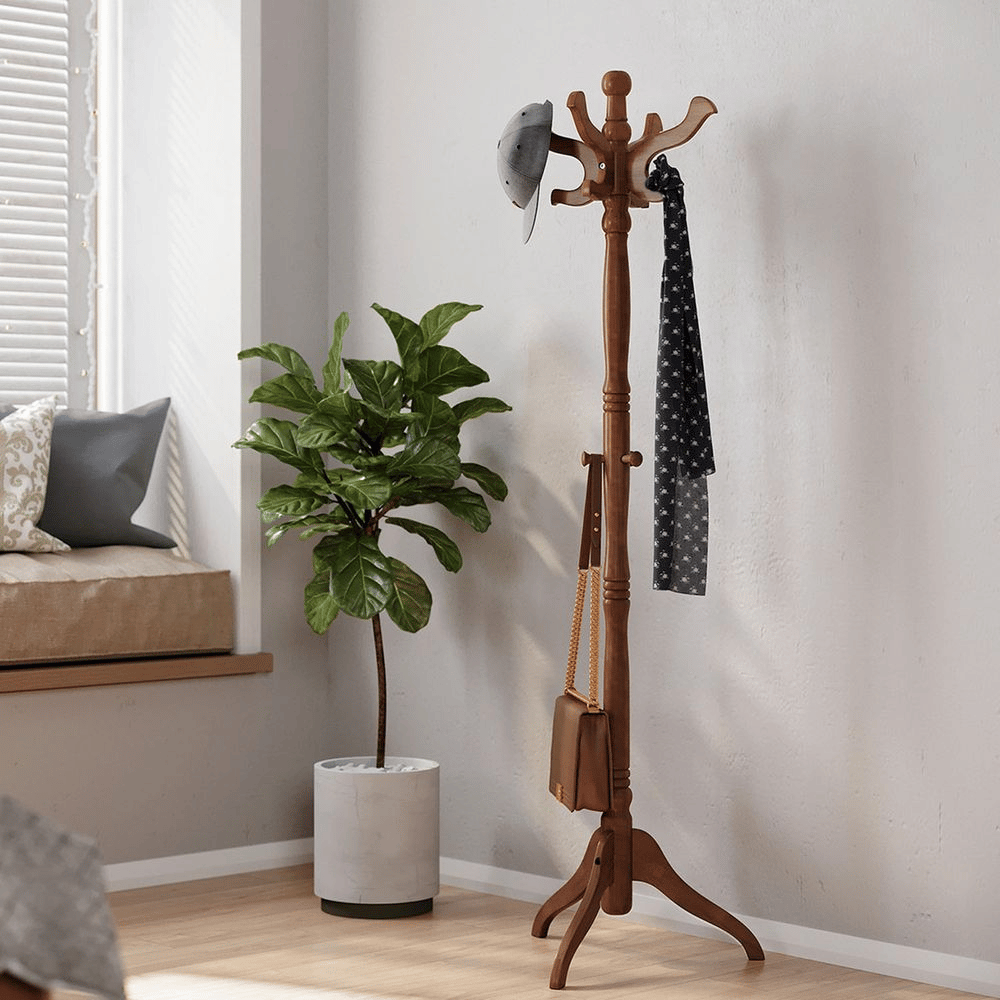 A wooden coat rack stands with a cap, scarf, and bag hanging on it, next to a green potted plant.