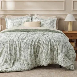 A bed set featuring a floral-patterned comforter and pillow shams in soft green tones, complemented by a neutral decorative pillow. A wooden nightstand is visible beside the bed with a lamp and small objects placed on it.