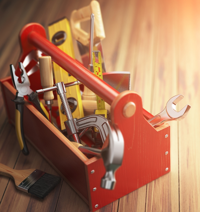 A red toolbox with various hand tools including a hammer, wrench, pliers, screwdrivers, and a paintbrush on a wooden surface.